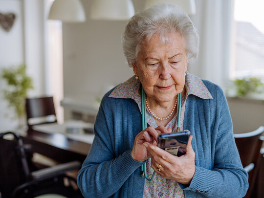 Eine ältere Frau steht in ihrem Esszimmer und bedient ein Smartphone. Sie tippt etwas ein.