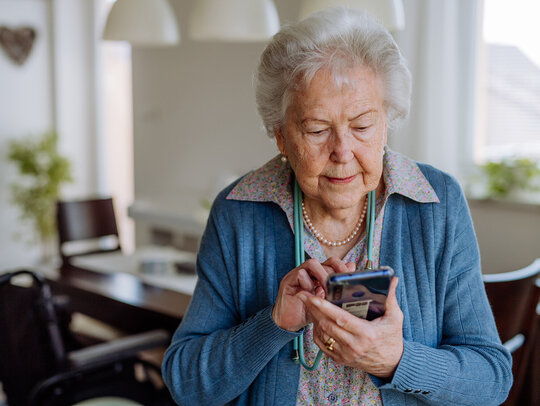 Eine ältere Frau steht in ihrem Esszimmer und bedient ein Smartphone. Sie tippt etwas ein.