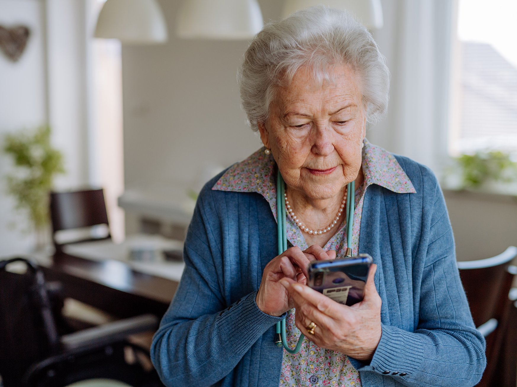 Eine ältere Frau steht in ihrem Esszimmer und bedient ein Smartphone. Sie tippt etwas ein.
