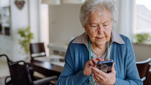 Eine ältere Frau steht in ihrem Esszimmer und bedient ein Smartphone. Sie tippt etwas ein.