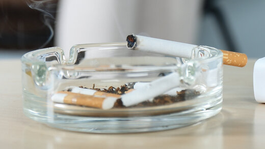 Ein voller Aschenbecher mit glimmender Zigarette steht auf einem Tisch.