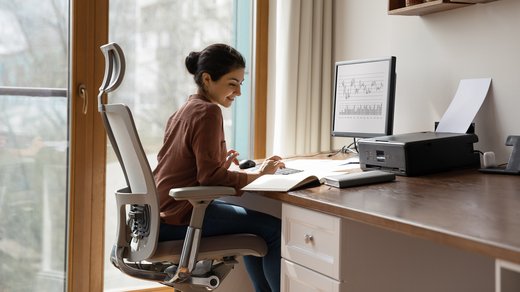 Eine Frau sitzt in einem aufgeräumt wirkenden Home Office an ihrem Arbeitsplatz. Die Möbel erscheinen ergonomisch und das Arbeitszimmer aufgeräumt. 