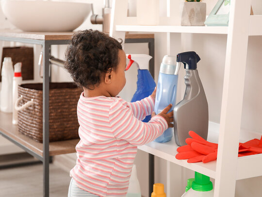 Ein Baby greift nach Reinigungsmitteln, die im untersten Fach eines Regals aufbewahrt werden. Die Situation ist potenziell gefährlich und weist auf Vergiftungsgefahren mit Reinigungsmitteln hin.