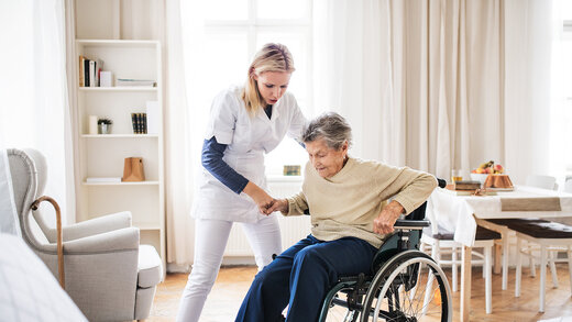 Eine Pflegerin unterstützt eine Seniorin im Rollstuhl beim Aufstehen; die Szene zeigt eine Situation der häuslichen Pflege.