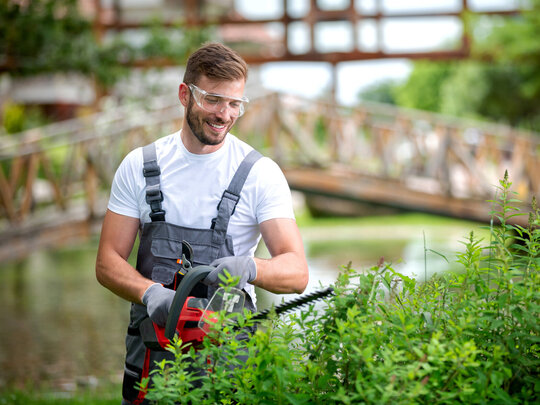 Mann schneidet Hecke mit elektrischem Heckenschneider und trägt Schutzbrille