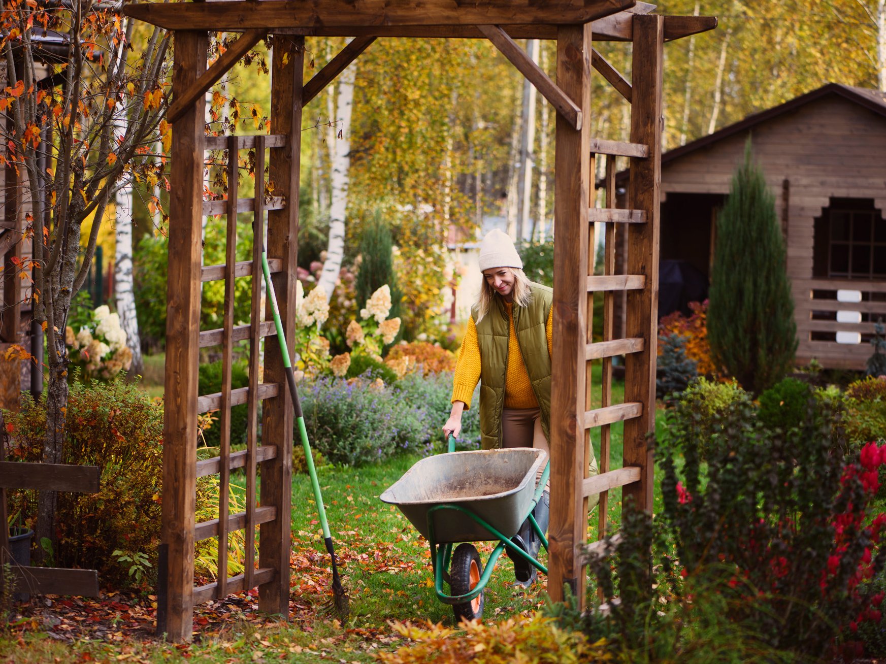 Eine Frau in Winterkleidung schiebt eine Schubkarre durch einen herbstlichen Garten.