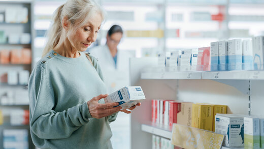 Eine ältere Frau mit weißen Haaren hält eine Medikamentenpackung in einer Apotheke. Sie liest die Beschreibung auf der Verpackung. 