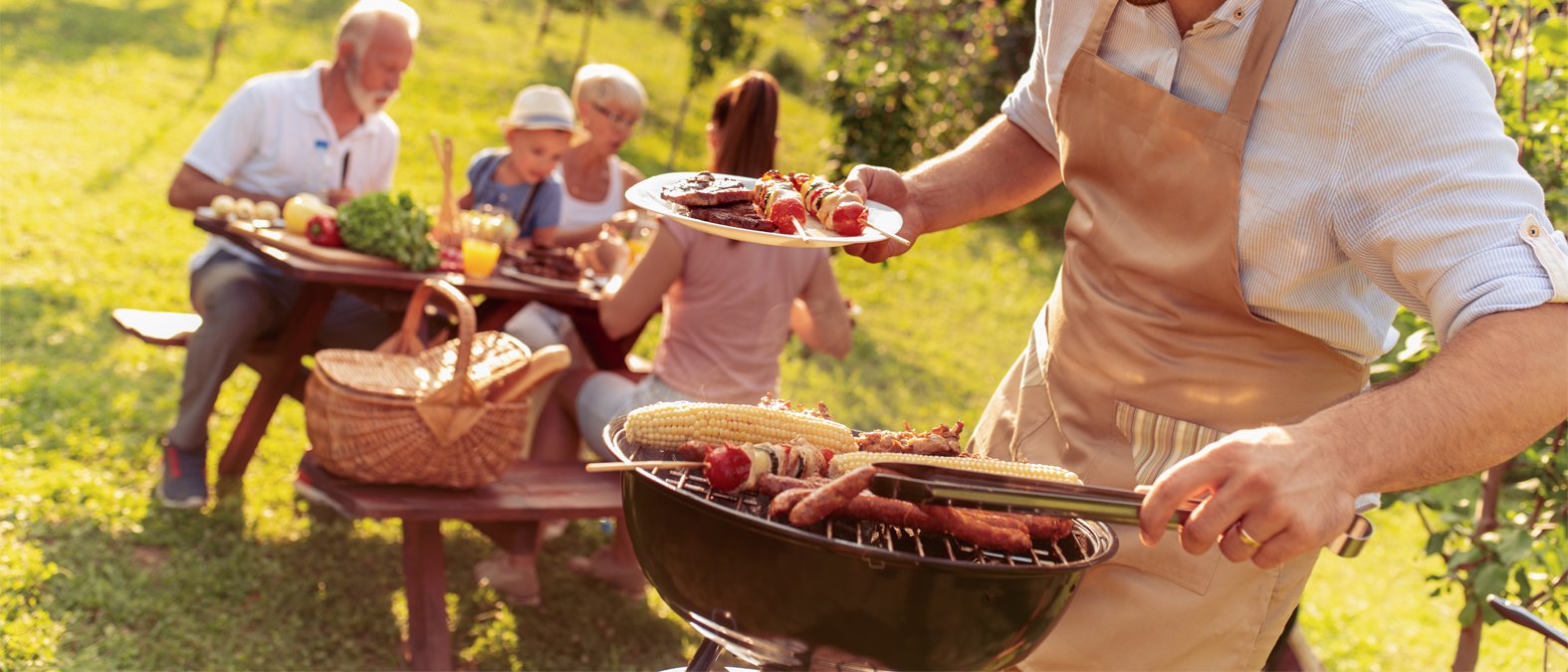 Eine Familie sitzt zusammen in einem Garten. Der Vater steht in einer hellbraunen Schürze am Grill und wendet das Grillgut. Die Stimmung scheint ausgelassen zu sein. 