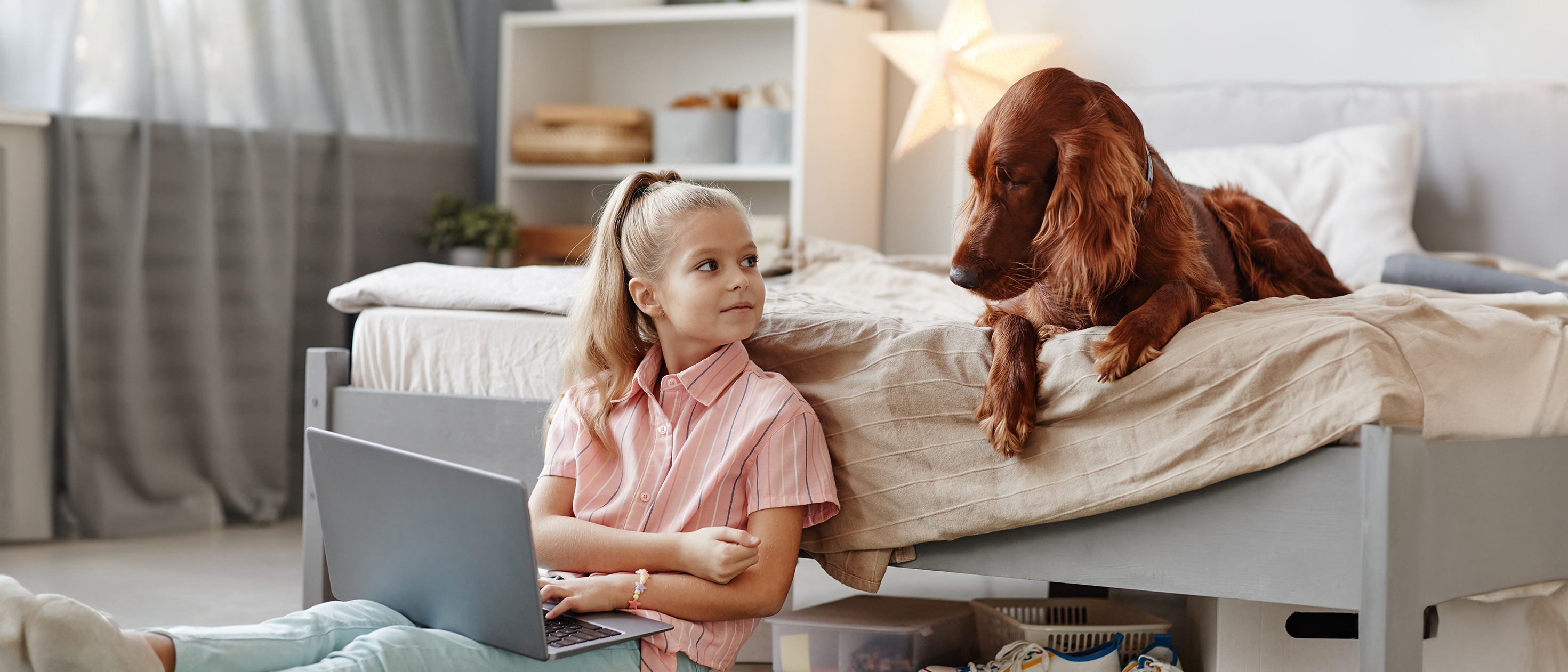 Ein Mädchen sitzt auf dem Boden vor ihrem Bett im Kinderzimmer. Auf dem Bett sitzt ein grosser brauner Hund und schaut aufmerksam in die Umgebung. 