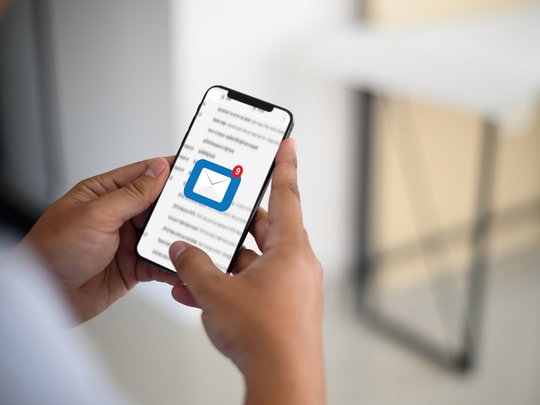 Foto eines Smartphones, in den Händen einer jungen Person. Das Display zeigt einen E-Mail-Posteingang.