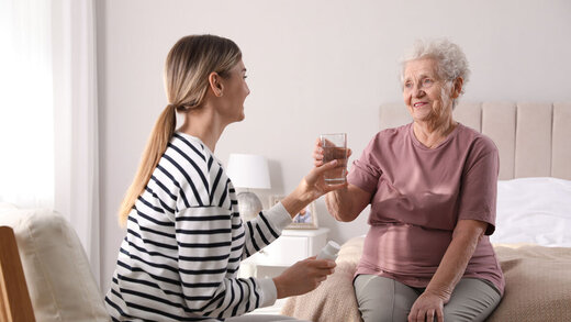 Junge Frau reicht einer älteren Dame ein Glas Wasser zur Erinnerung ans Trinken.