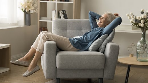 Frau entspannt in ihrem Wohnzimmer, sitzt in einem Sessel und verschränkt die Arme hinter dem Kopf. Der Raum vermittelt eine ruhige, komfortable Atmosphäre, geeignet für Erholung und Entspannung.
