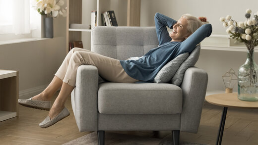 Frau entspannt in ihrem Wohnzimmer, sitzt in einem Sessel und verschränkt die Arme hinter dem Kopf. Der Raum vermittelt eine ruhige, komfortable Atmosphäre, geeignet für Erholung und Entspannung.