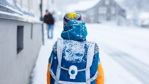 Ein Schulkind befindet sich im Winter auf dem Schulweg und geht die Straße entlang. Die Straßen sind stark mit Schnee bedeckt.