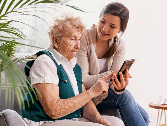 Eine junge Frau erklärt einer Senioren, wie sie ihr Smartphone bedienen kann. Die Seniorin wirkt konzentriert. 