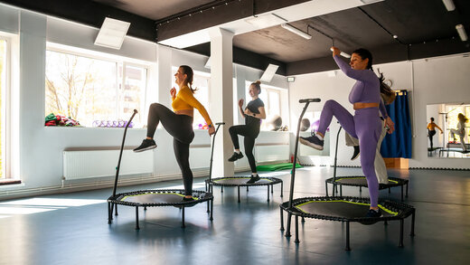 Eine Gruppe von Menschen macht in einem großen Raum Fitness auf Indoor-Trampolinen. Sie tragen alle Sportkleidung. 