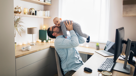 Ein Mann sitzt im Home Office an seinem Schreibtisch. Er hebt ein Baby in die Luft und küsst es auf die Wange. 