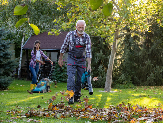 Ein Senior in Latzhose läuft mit einem Laubbläser durch den Garten. Im Hintergrund bedient eine junge Frau einen Rasenmäher.
