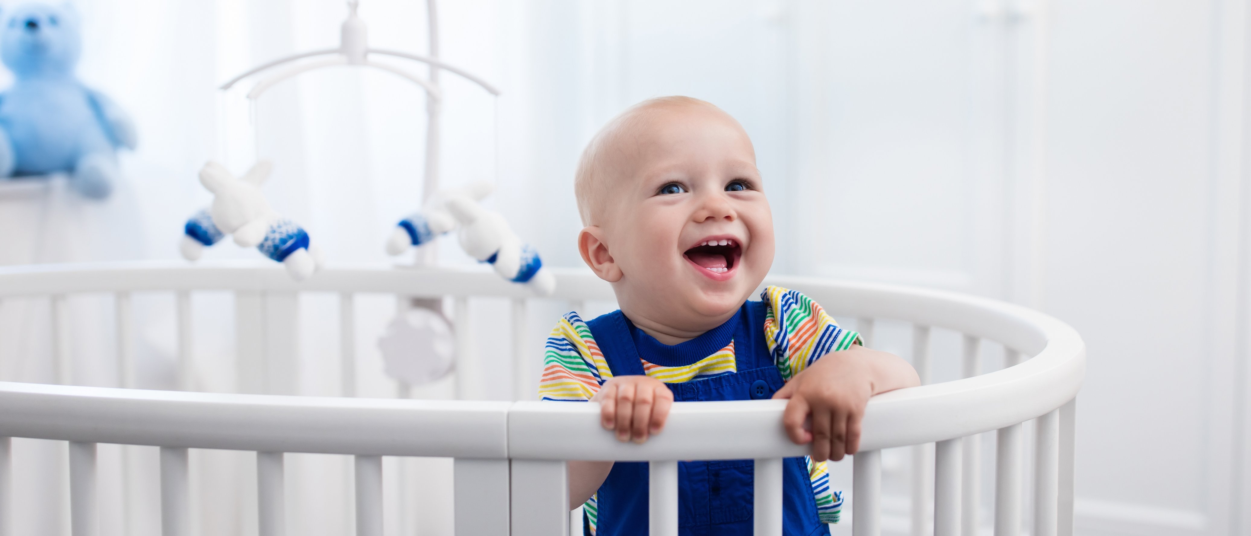 Ein Baby liegt lachend in einem Kinderbett, hält sich an den Gitterstäben fest und schaut fröhlich nach vorne. 