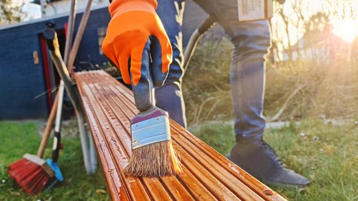 Jemand pinselt Leinöl auf ein Stück Holz. Die Person befindet sich in einem Garten. 