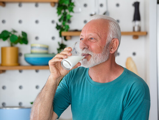 Ein Senior steht in seiner Küche und trinkt ein Glas Milch.