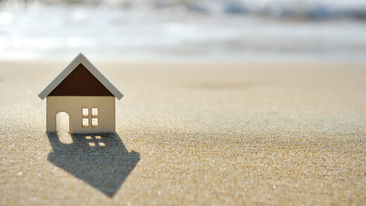 Ein kleines Häuschen auf Holz steht im Sand vor einem Meer. 