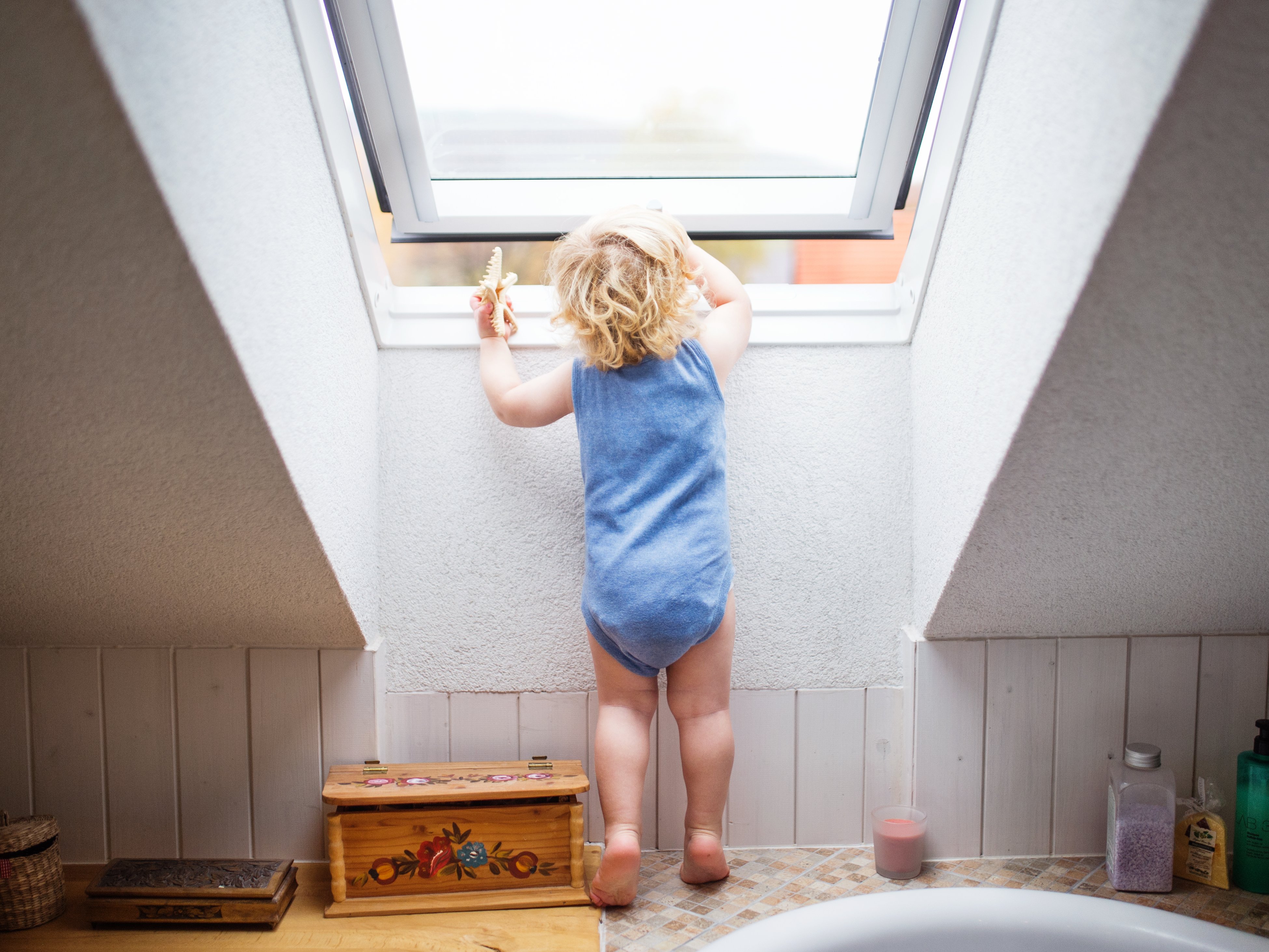 Kleinkind steht an einem halb geöffneten Dachfenster im Bad und schaut neugierig nach draußen.