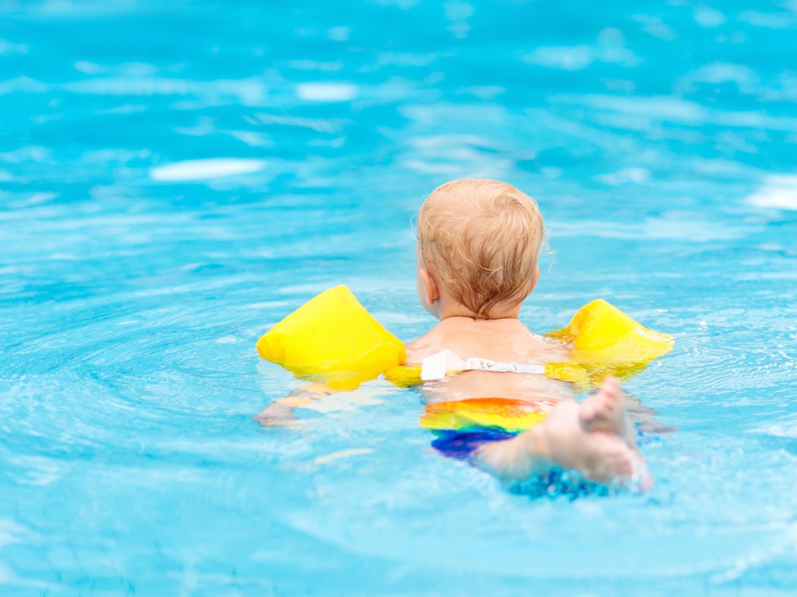 Ein Kleinkind ist von hinten im Pool zu sehen, mit gelben Schwimmflügeln.