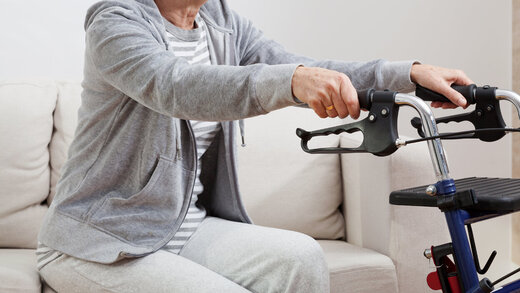 Eine Person hält sich an einem Rollator fest und stützt sich darauf ab, um vom Sofa aufzustehen. 