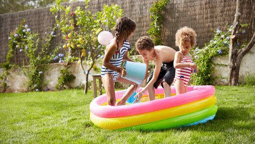Drei Kinder planschen fröhlich in einem kleinen aufblasbaren Schwimmbecken im Garten. Wasser spritzt in die Luft. 