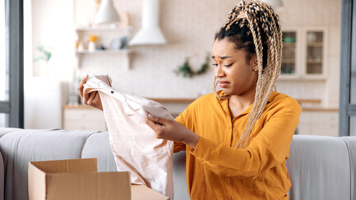 Eine junge Frau sitzt in ihrem Wohnzimmer und packt ein Paket aus. Sie hält ein Kleidungsstück in ihren Händen und schaut skeptisch. 