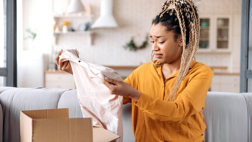 Eine junge Frau sitzt in ihrem Wohnzimmer und packt ein Paket aus. Sie hält ein Kleidungsstück in ihren Händen und schaut skeptisch. 