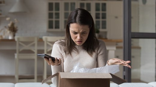 Eine junge Frau schaut verärgert in einen Karton, der geöffnet auf ihrem Schoß steht. Sie scheint mit ihrer Bestellung nicht zufrieden zu sein.