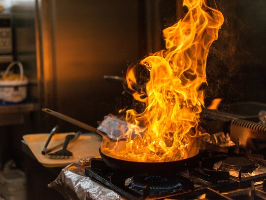 „Fettbrand in einer Pfanne. Die Szene zeigt eine gefährliche Situation in der Küche, in der heißes Fett Feuer fängt, was zu einer potenziellen Brandgefahr führen kann. Es wird auf die Bedeutung der richtigen Handhabung von Fett und heißen Ölen hingewiesen, um Unfälle zu vermeiden.