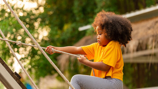 Ein Mädchen mit dunklen Locken und bunter Kleidung klettert auf dem Klettergerüst eines öffentlichen Spielplatzes. 