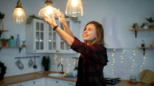 Junge Frau wechselt eine Glühbirne in der Küche. Beim Arbeiten mit elektrischen Geräten besteht die Gefahr eines Stromschlags.