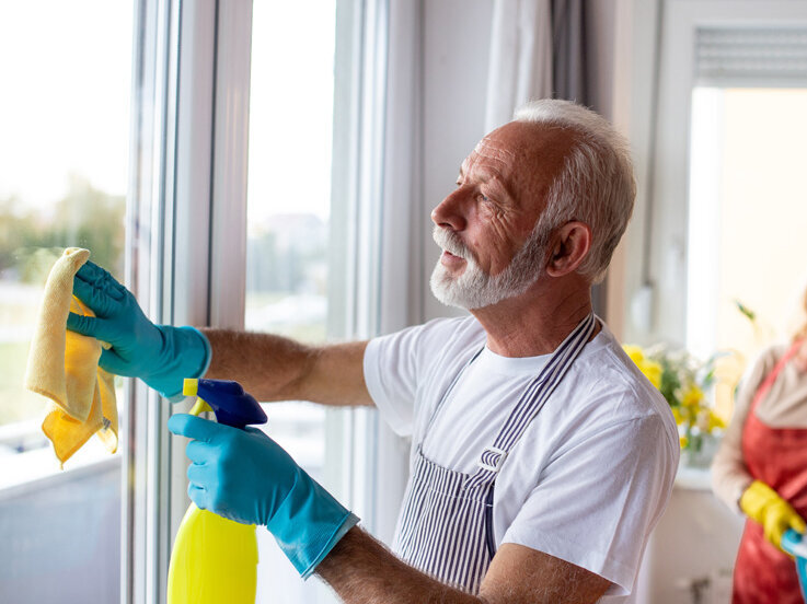 Ein älteres Paar putzt gemeinsam die Wohnung. Die Frau schaut zu, während ihr Mann die Fenster putzt. 