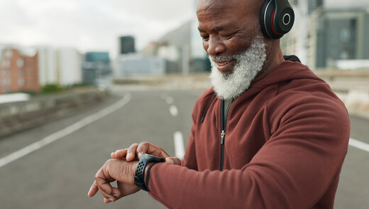 Ein Senior in Sportkleidung und mit Kopfhörern schaut etwas auf seiner Smartwatch nach. Er steht auf einer Straße. 