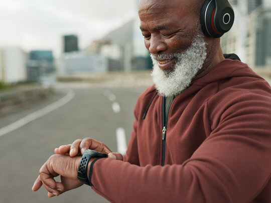 Ein Senior in Sportkleidung und mit Kopfhörern schaut etwas auf seiner Smartwatch nach. Er steht auf einer Straße.