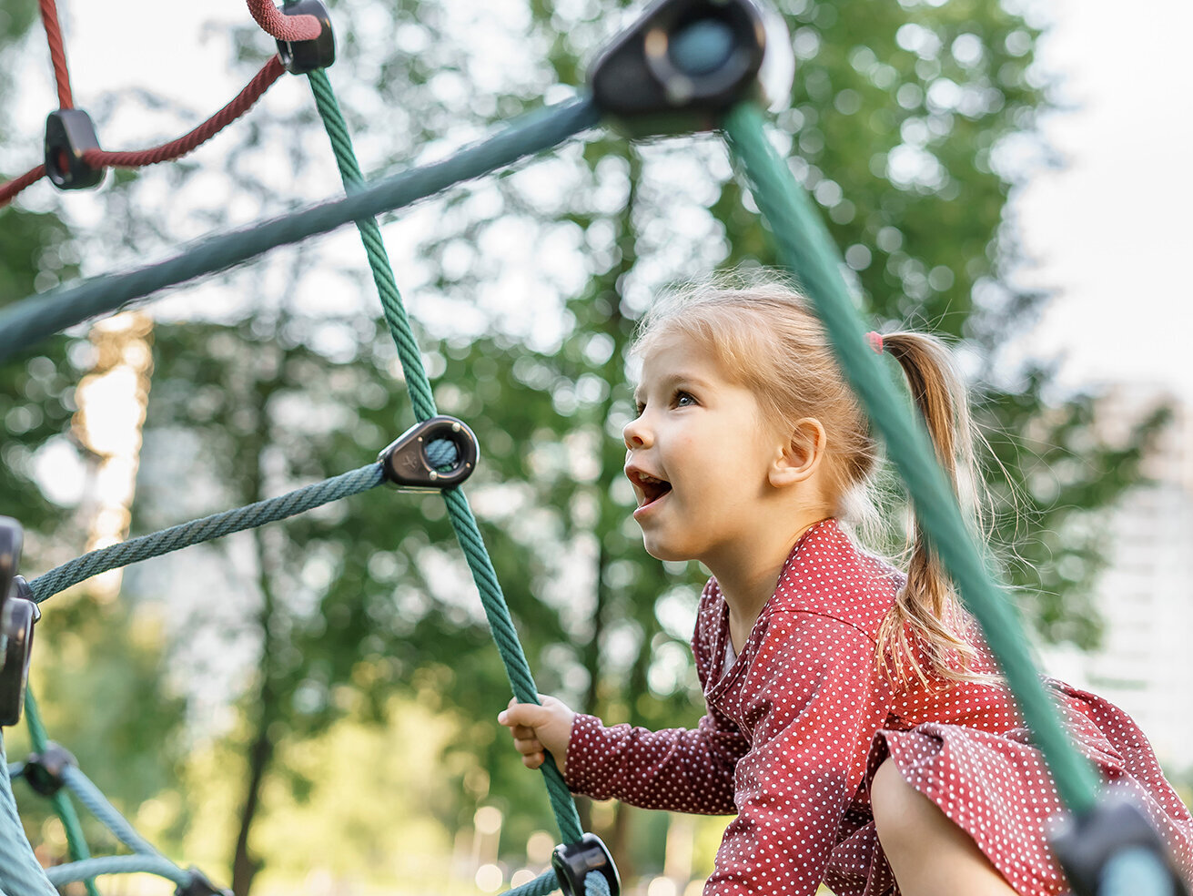 Ein kleines Mädchen in einem Kleid klettert auf dem Klettergerüst eines öffentliches Spielplatzes.