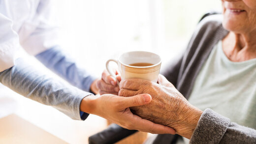 Eine pflegende Person reicht einer Senioren eine Tasse, um sie ans Trinken zu erinnern. 