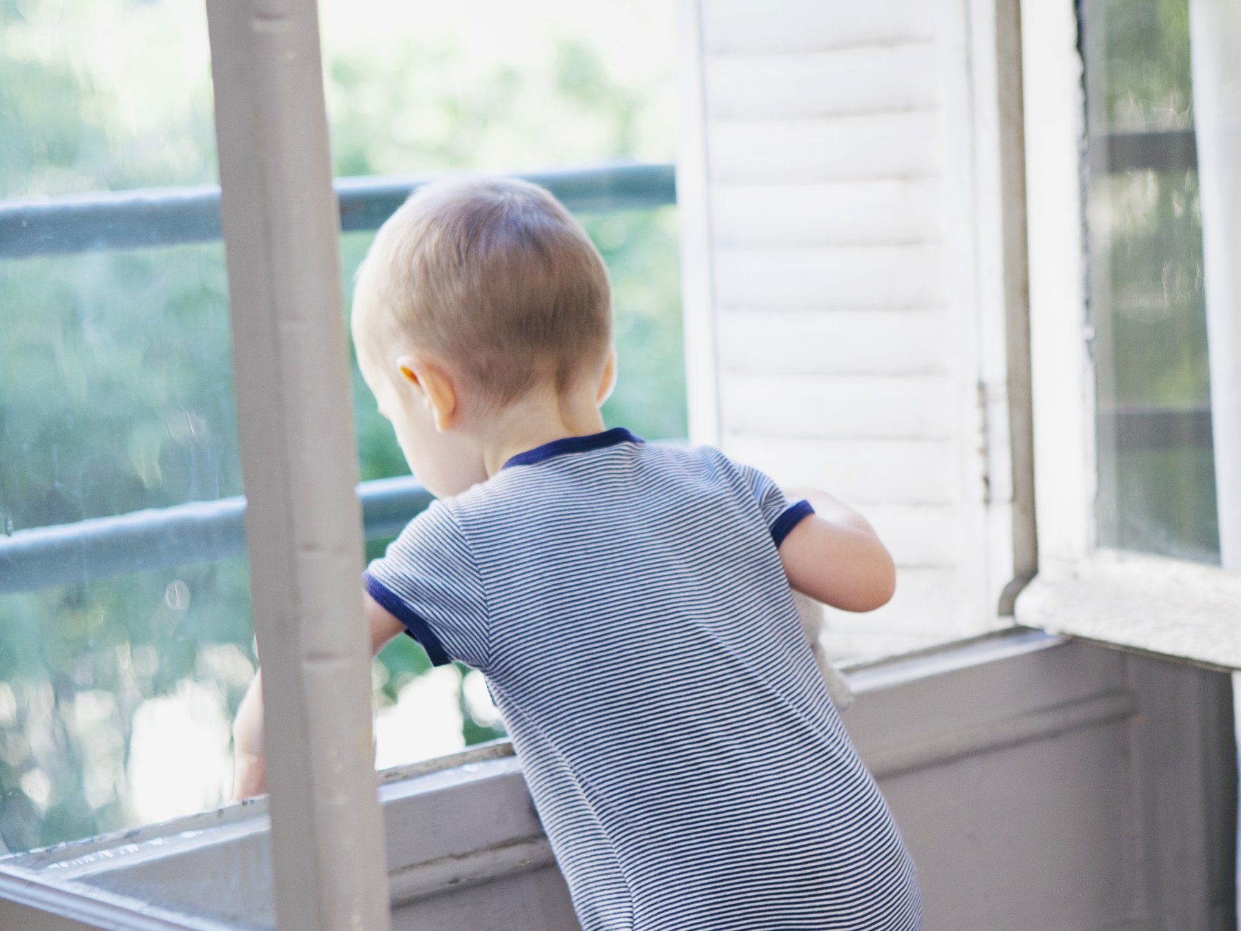 Kleinkind steht an einem geöffneten Fenster und streckt die Arme hinaus. Das Bild zeigt eine gefährliche Situation.