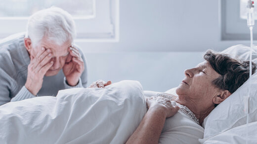 Älterer Herr sitzt am Krankenbett seiner Frau und stützt seinen Kopf auf seine Hände