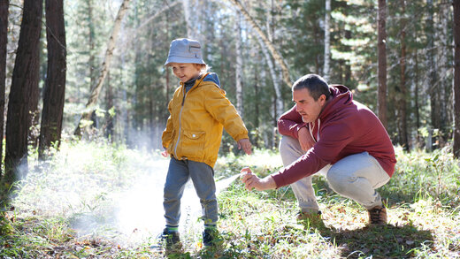 Ein Vater befindet sich mit seinem kleinen Sohn in einem Wald und sprüht ihn mit Zeckenschutzspray ein.